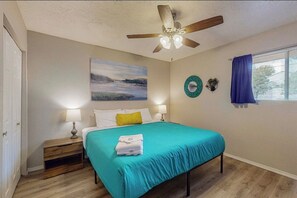 Un dormitorio con cama, dos mesitas de noche, ventilador de techo, ventana con cortinas y una obra de arte colgada en la pared.