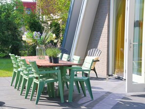 An outdoor patio with a wooden table and green chairs, surrounded by potted plants and a glass door. - Villa in Callantsoog With Swimming Pool (Callantsoog)