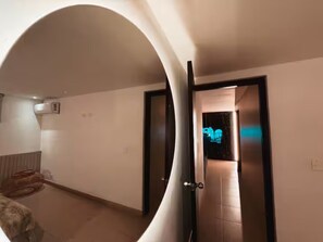A hallway with a curved mirror reflecting a room, a door with a neon sign, and a small rug on the floor. - Oasis Loft Friends (Medellín)