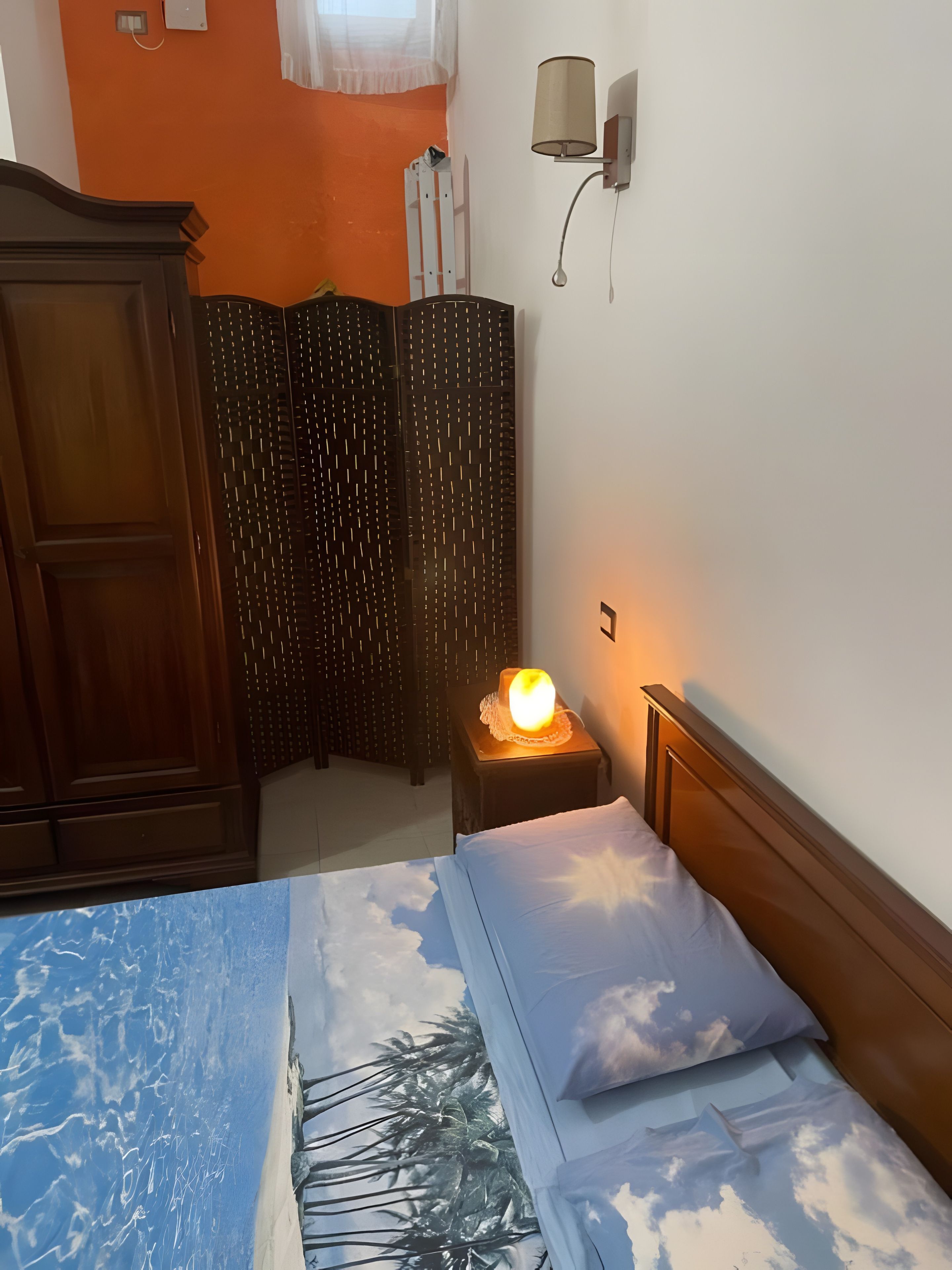 Un dormitorio con una cama que presenta un diseño con cielo y nubes, un armario de madera, una mesita de noche con lámpara y un aplique de pared.