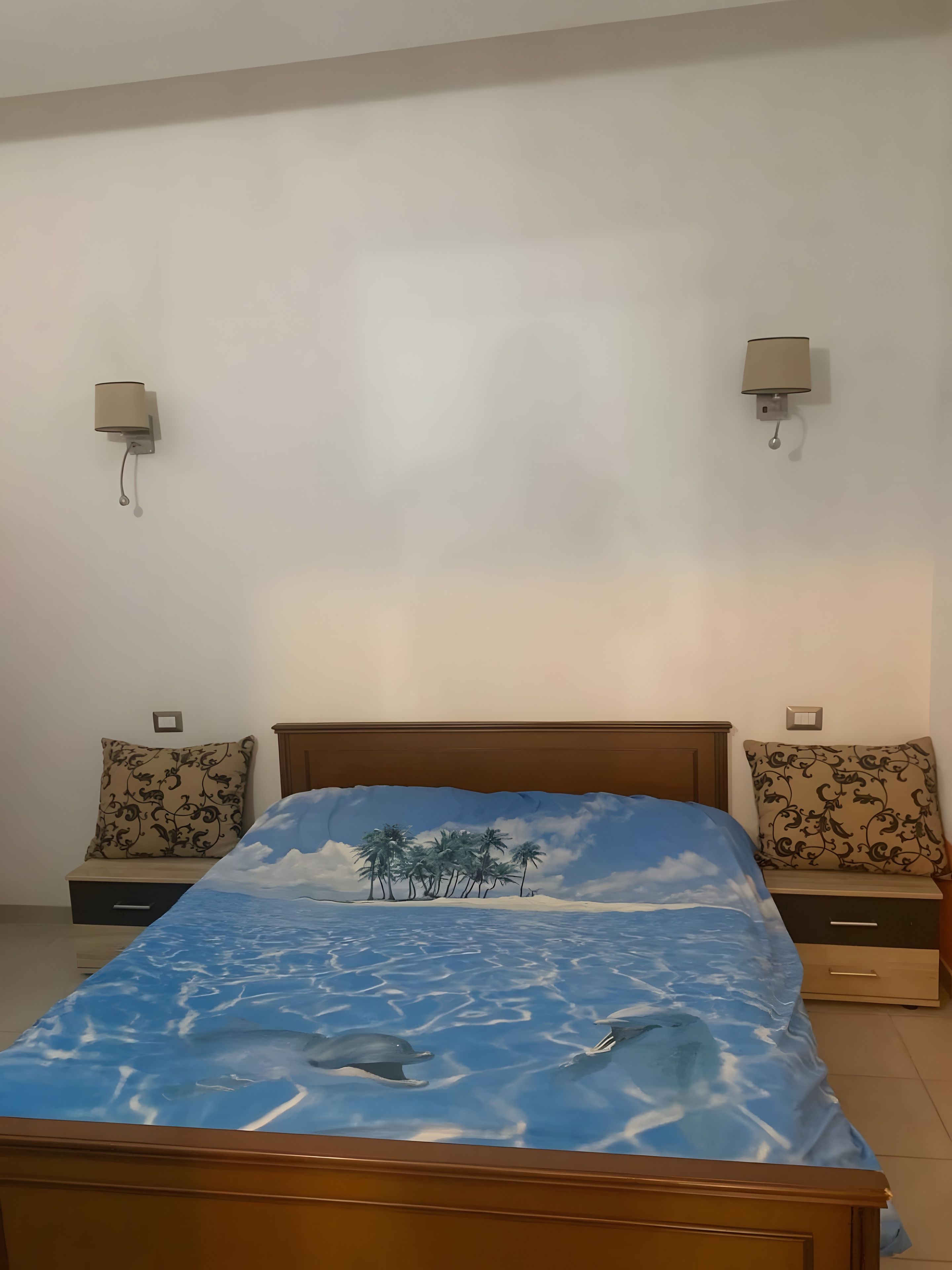 Een slaapkamer met een bed met een tropisch stranddesign, twee nachtkastjes met lampen en een houten hoofdbord.