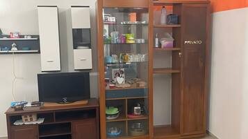 A room with a TV on a wooden stand, a wardrobe with glass doors, and a shelf with various items.