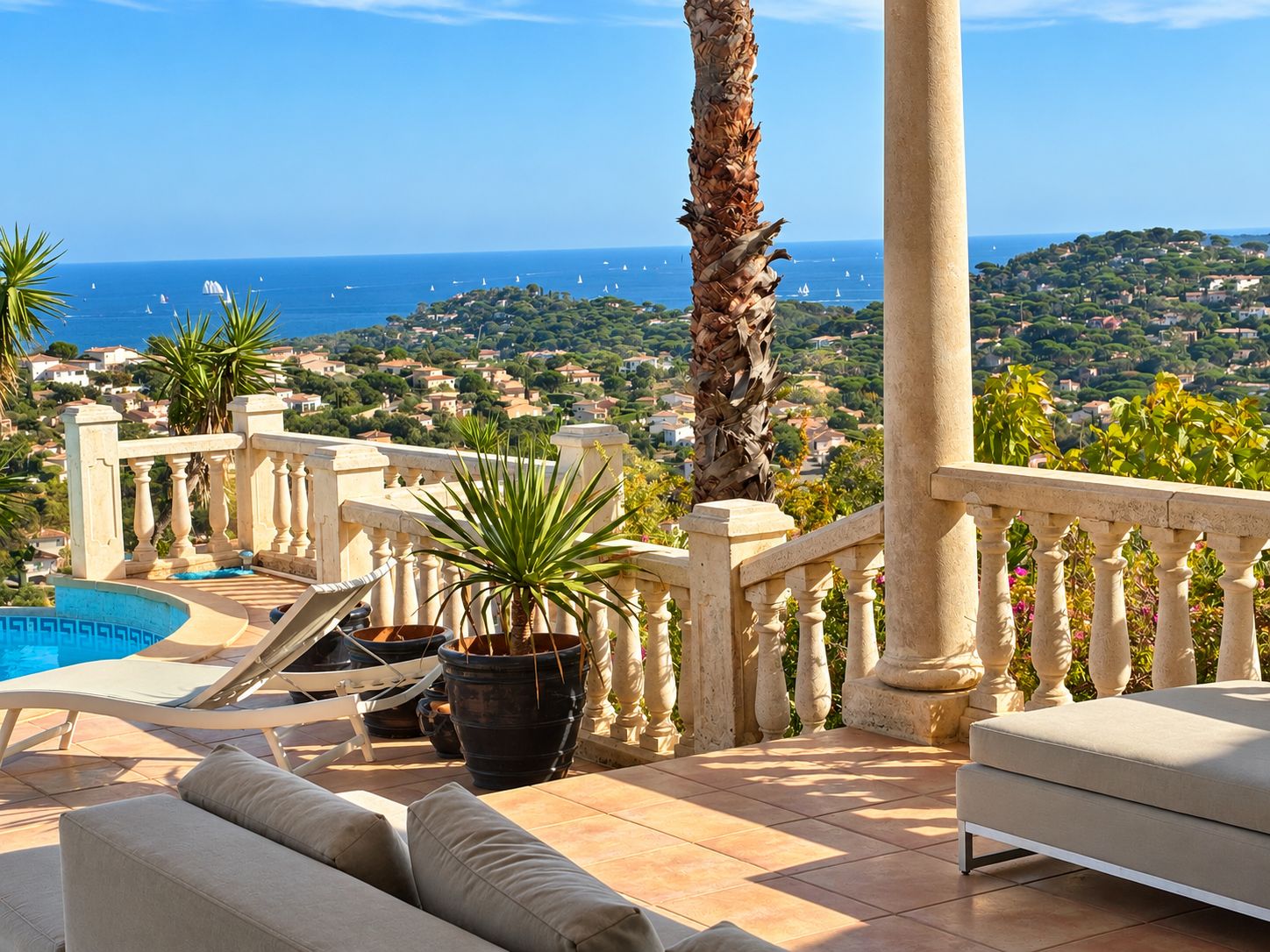 A terrace with a pool, lounge chairs, and a view of the sea and a town.