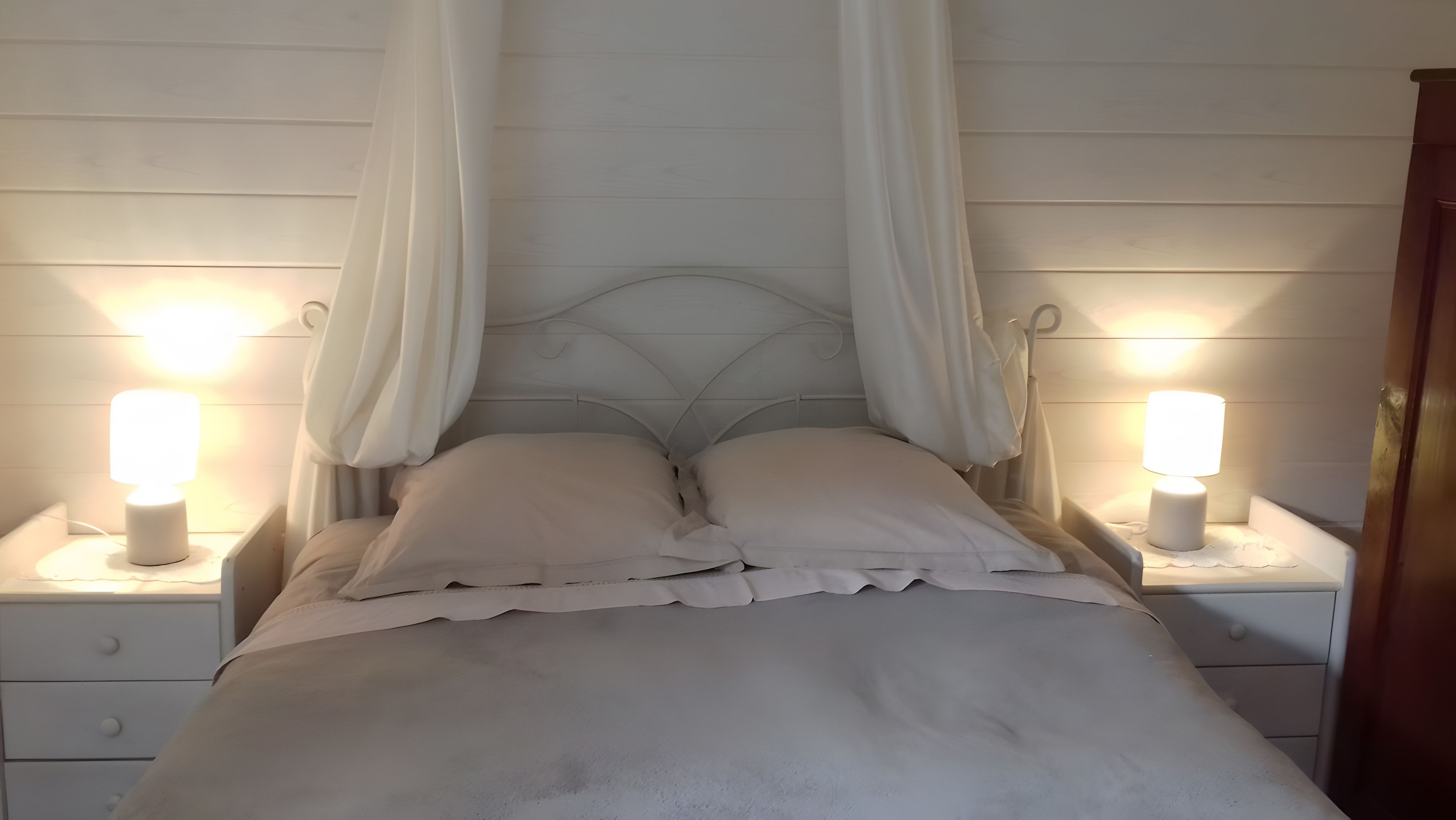 En säng med vita sängkläder och ett himmelstäng, med sänglampor på nattduksborden på var sin sida.