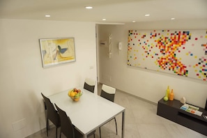 A dining area with a white table, black chairs, and a fruit bowl. There is a colorful pixelated artwork on the wall and a door leading to another room. - Dante House (Bari)
