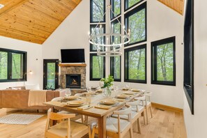 Un comedor con una larga mesa de madera dispuesta para una comida, rodeada de sillas, con una chimenea y ventanas grandes al fondo.