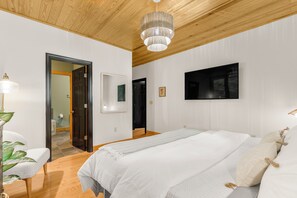 Un dormitorio moderno con una cama grande, techo de madera, televisor montado y una puerta que da a otra habitación.