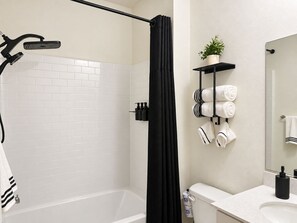 A bathroom with a white bathtub, a black shower curtain, a toilet, a sink, and a mirror. - Modern Organic SoCal Retreat - 2BR + 2.5BA + GYM (Placentia)