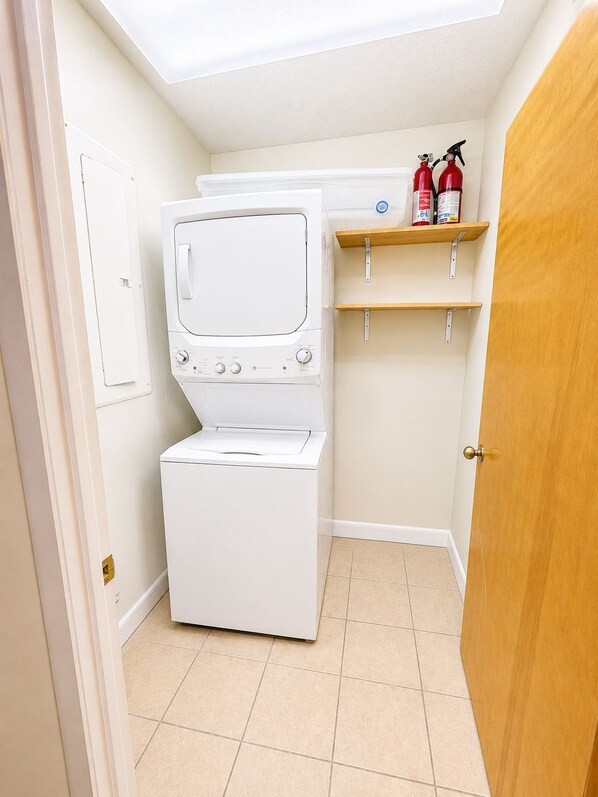A laundry room with a stacked washer and dryer, a shelf with fire extinguishers, and a door leading to another room. - Beech Haven | Ski-In/Out • Sauna • Sleeps 18 (Beech Mountain)