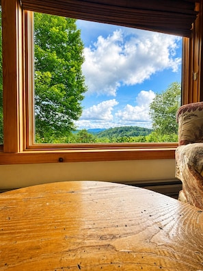 A room with a large window offering a view of a forest and hills. - Tranquility House | Hot Tub • Views • Fireplace (Banner Elk)