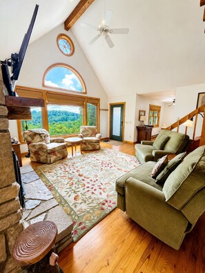 A spacious living room with a high ceiling, a fireplace, a large window offering a view of the outdoors, and a cozy seating area. - Tranquility House | Hot Tub • Views • Fireplace (Banner Elk)