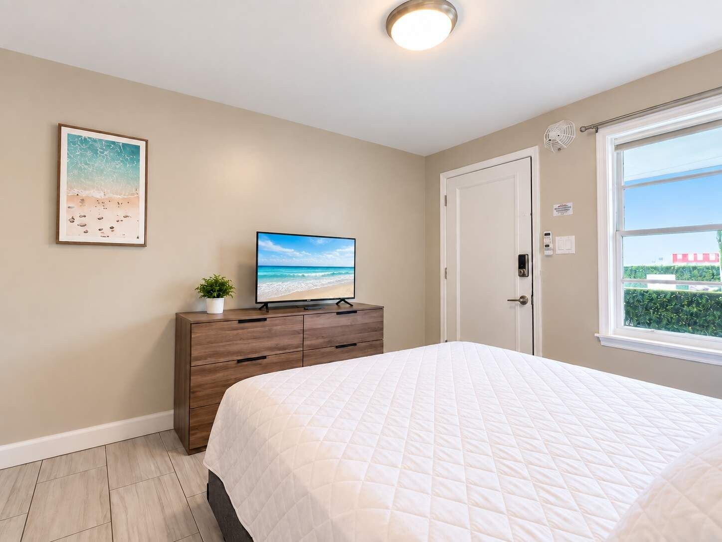 Een slaapkamer met een bed, een televisie op een houten dressoir, een schilderij van een strand aan de muur en een raam met uitzicht op groen.
