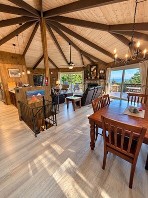 A spacious dining area with a wooden table and chairs, a kitchen in the background, and a large window offering a view of the outdoors. - Beech Nutz - mountain view, deck and wood burning fireplace (Beech Mountain)