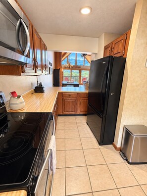 A kitchen with wooden cabinets, a black refrigerator, a stove, and a view of the outdoors through large windows. - Ski Side Retreat - Walk to Slopes, Gas Fireplace, Close to Town (Beech Mountain)