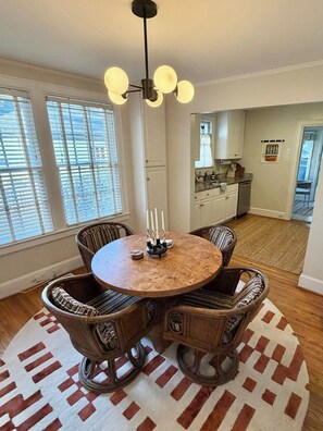 A dining area with a round table surrounded by wicker chairs, a chandelier, and a view of the kitchen. - Charming Historic Bungalow · Walkable to Eats/Park (Charlotte)