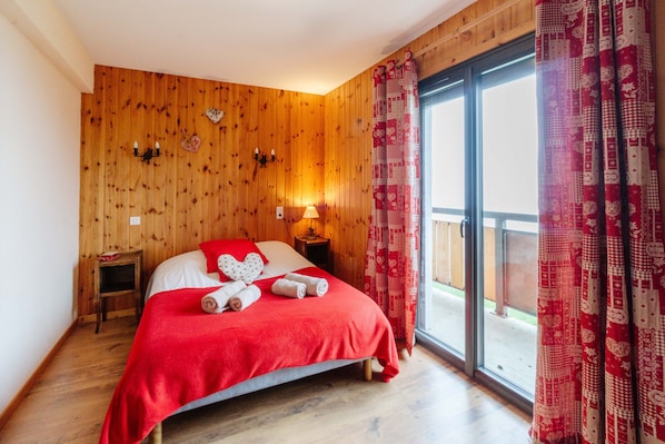 A bedroom with a wooden wall, a bed with a red blanket, two bedside tables, a nightstand, and a window with patterned curtains. - Apartment 'Sabots De Vénus' with Mountain View and Balcony (Fontcouverte-la-Toussuire)