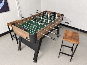 Ein Kicker mit Holzrahmen und schwarzen Beinen, ein kleiner Hocker und eine weiße Wand mit einem Gemälde im Hintergrund.