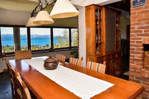 A dining area with a long wooden table, two hanging lamps, and a view of a lake through large windows. - Casa cerca del centro para 6 con vista al lago (San Carlos de Bariloche)