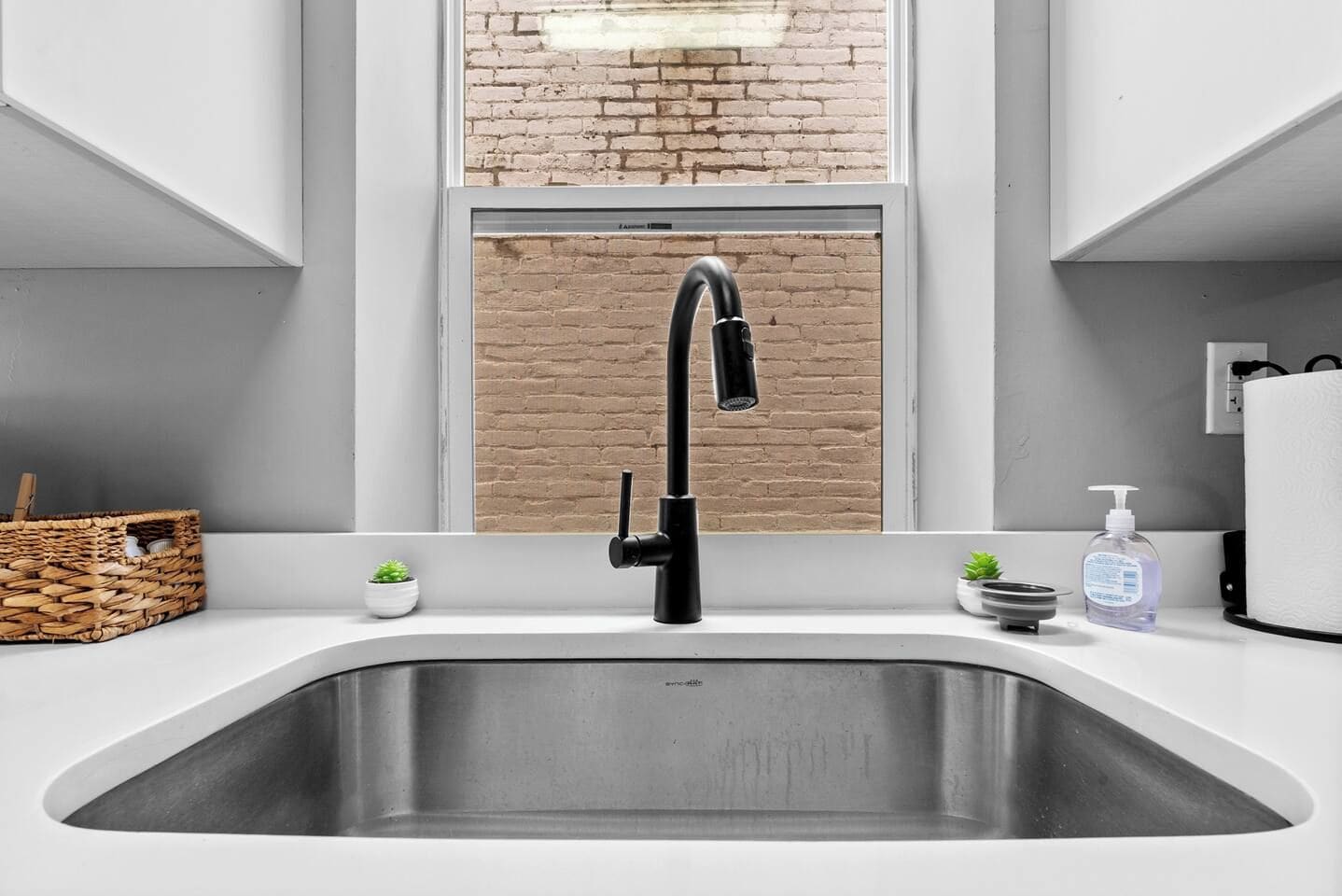Una cocina con un grifo moderno, una ventana con vista a un muro de ladrillo, una pequeña planta en maceta y un dispensador de jabón.