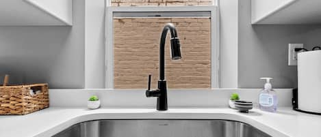 Una cocina con un grifo moderno, una ventana con vista a un muro de ladrillo, una pequeña planta en maceta y un dispensador de jabón.