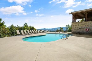 A swimming pool with a stone wall and lounge chairs, surrounded by trees and a view of mountains. - STUNNING Mountain Views! Sleeps 10, hot tub, game room, 3 Panoramic Decks (Sevierville)