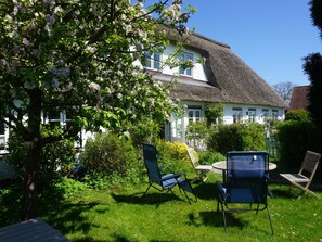 Ein Haus mit Strohdach, ein Garten mit blühendem Baum und Gartenmöbeln.