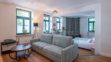 A modern living room with a sofa, a small table, and a floor lamp. There is a bed with a white mattress and a window with curtains.