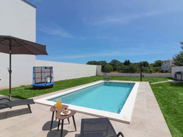 Ein rechteckiger Swimmingpool mit einer gefliesten Liegefläche, ein kleiner Tisch mit Getränken und Speisen, ein blauer Trampolin und ein überdachter Sitzbereich.