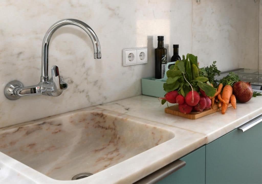 Una cocina con un lavabo, grifo y encimera. Hay botellas y verduras sobre la encimera.