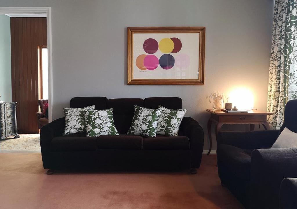 A living room with a dark-colored sofa, two patterned throw pillows, a framed abstract artwork on the wall, and a side table with a lamp.