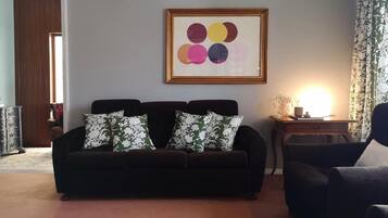 A living room with a dark-colored sofa, two patterned throw pillows, a framed abstract artwork on the wall, and a side table with a lamp.