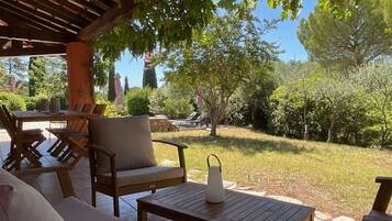 An outdoor patio area with wooden furniture, a stone table, and a view of a garden with trees and a lawn.