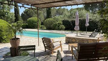A covered outdoor area with a pool, stone flooring, wooden furniture, and umbrellas.