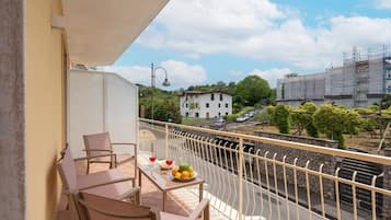 A balcony with a table set for two, a chair, and a view of a street with buildings and greenery.