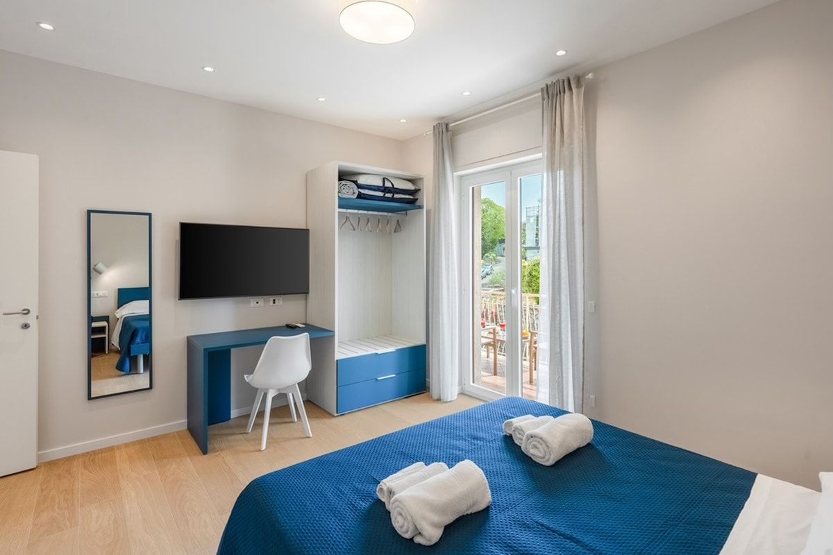 A hotel room with a bed, a desk, a television, a wardrobe, and a balcony with curtains.