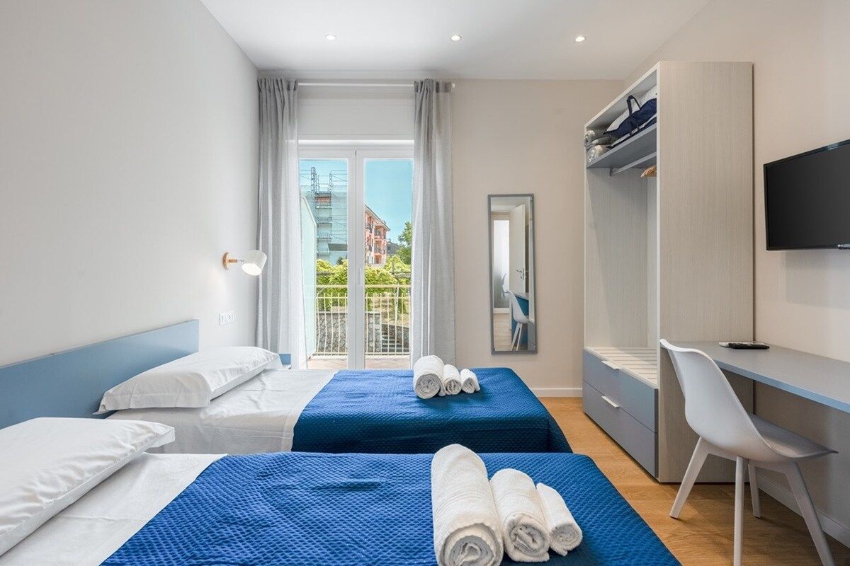 Ein Hotelzimmer mit zwei Betten, einem Schreibtisch, einem Stuhl, einem Fernseher und einem Balkon mit Blick auf Gebäude.