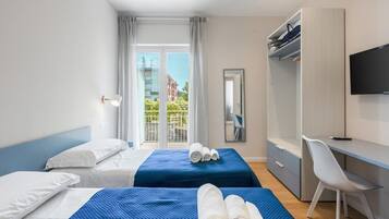 Ein Hotelzimmer mit zwei Betten, einem Schreibtisch, einem Stuhl, einem Fernseher und einem Balkon mit Blick auf Gebäude.