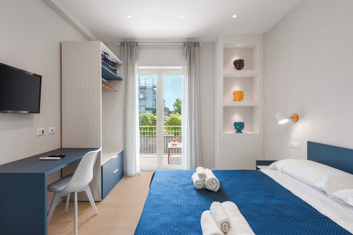 A hotel room with a bed, a desk, a chair, a television, and a balcony with a view of greenery.