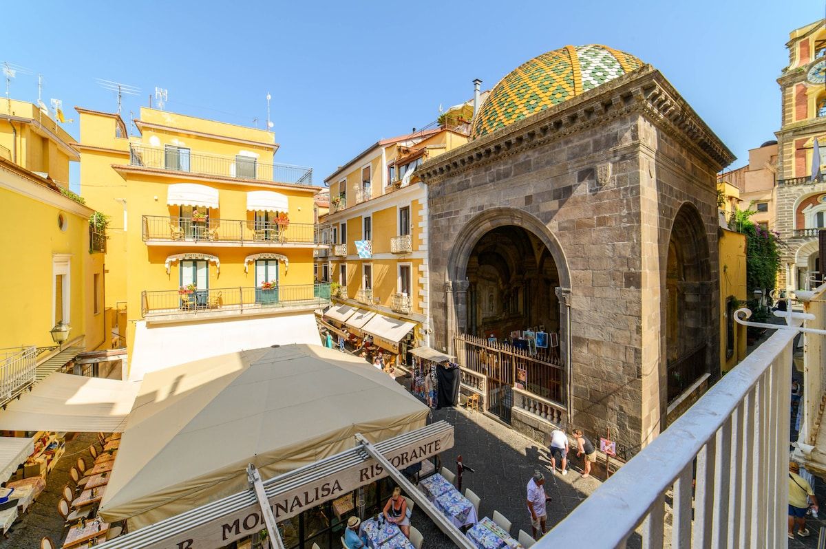A square with a stone archway, yellow buildings, and outdoor seating.