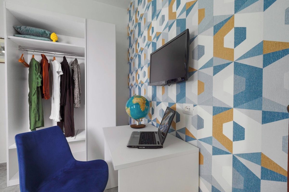Una habitación con un escritorio blanco, una computadora portátil, un globo terráqueo, un televisor en la pared y un perchero con varias prendas de vestir.