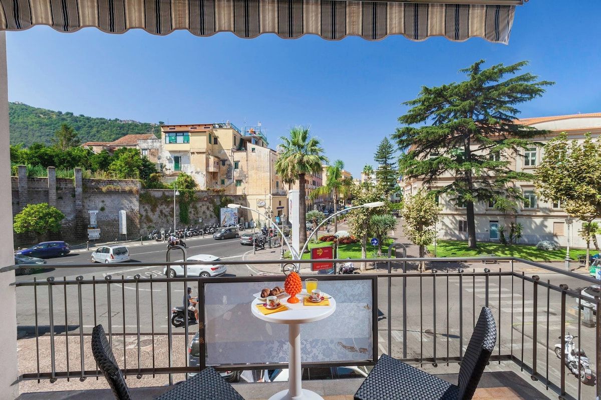 Un balcon avec une table préparée pour le petit-déjeuner, donnant sur une rue animée par la circulation des voitures et bordée d’immeubles.