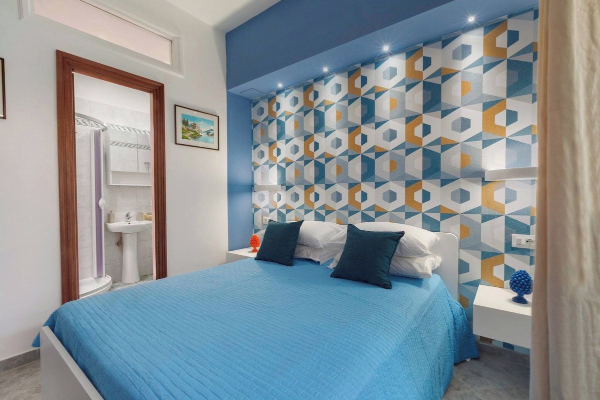 Une chambre à coucher avec un lit, une table de chevet, une étagère fixée au mur et une salle de bain visible à travers une porte ouverte.