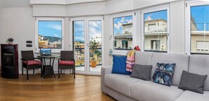 A living room with a sofa, chairs, a small table, and a TV. There are large windows with a view of buildings and a balcony. - Maya Penthouse - Sorrento Center (Sorrento)