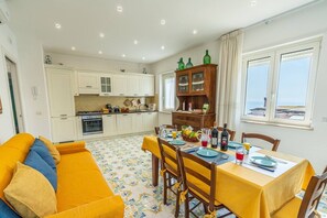 Cozinha compacta com armários brancos, uma mesa de jantar posta para dois e um sofá amarelo em primeiro plano.