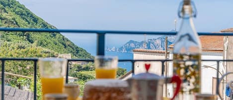 A table with a bottle, glasses, and a plate of food, overlooking a mountainous landscape and the sea.