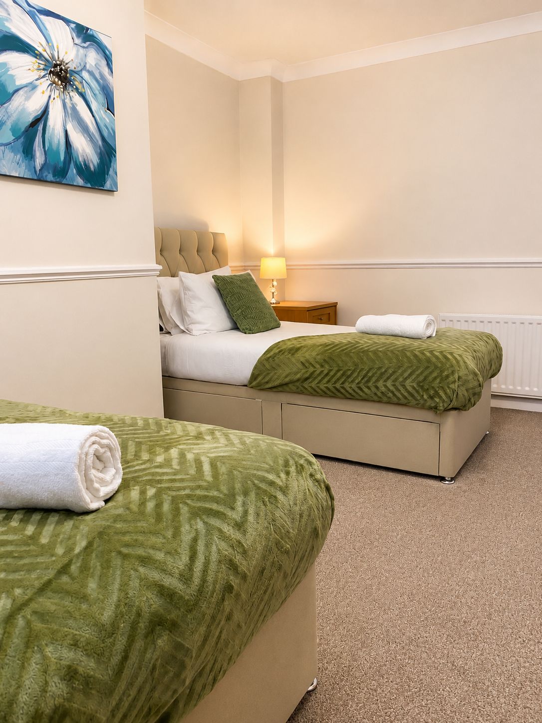A hotel room with two beds, a green patterned bedspread, a white towel, a lamp, and a painting on the wall.