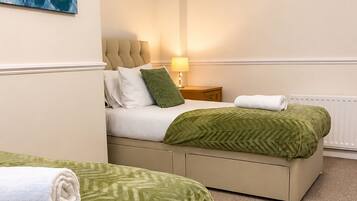 A hotel room with two beds, a green patterned bedspread, a white towel, a lamp, and a painting on the wall.