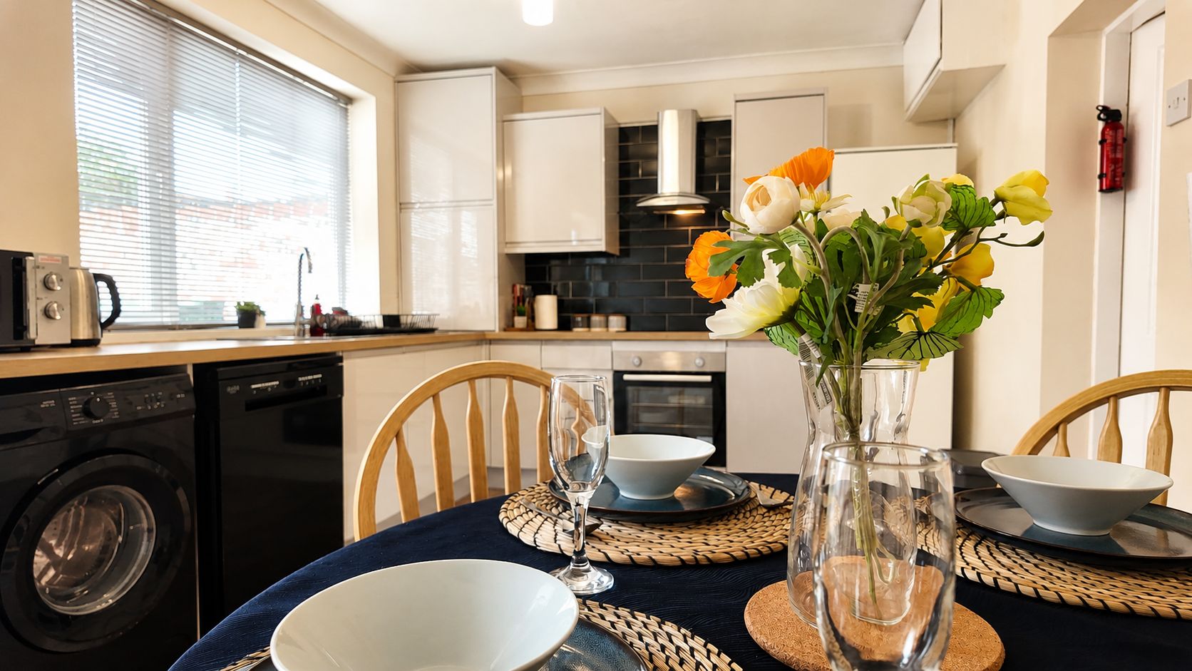 A modern kitchen with a dining table set for two, featuring a vase with flowers and a glass of water.