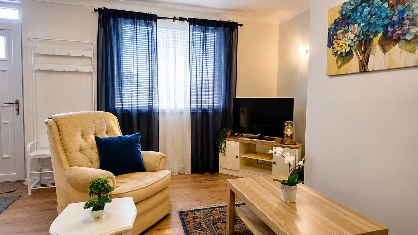A living room with a beige sofa, a coffee table, a TV stand with a flat-screen TV, a small plant, and a painting on the wall.