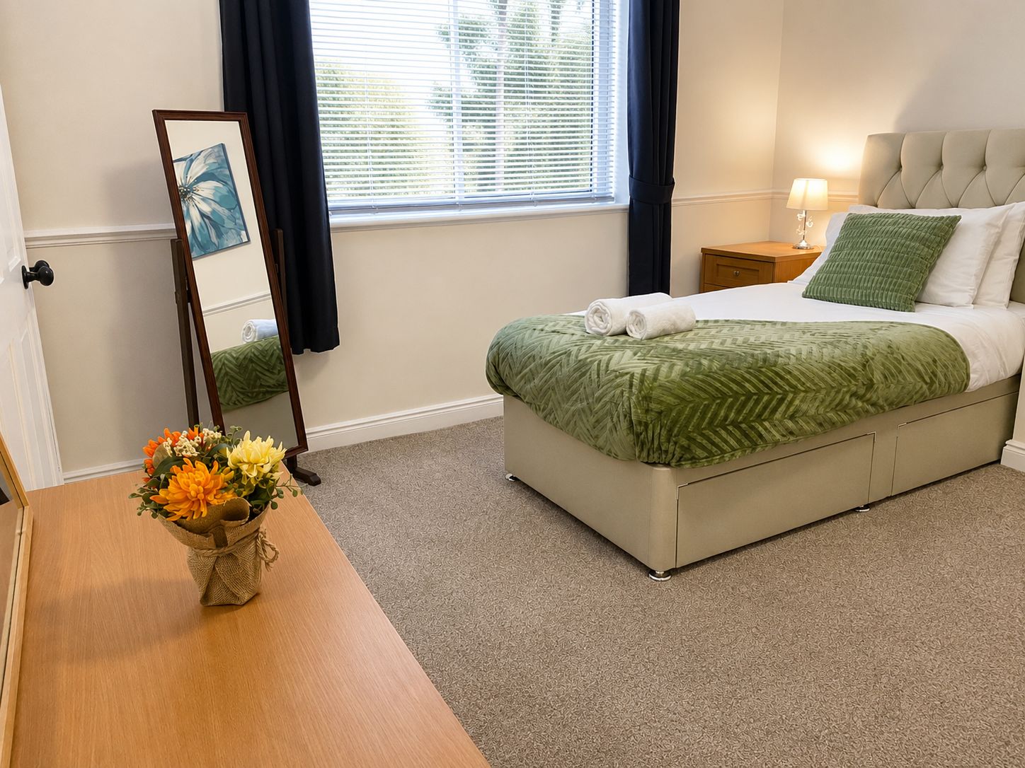 A neatly arranged bedroom with a bed, a bedside table, a lamp, a mirror, and a vase with flowers.